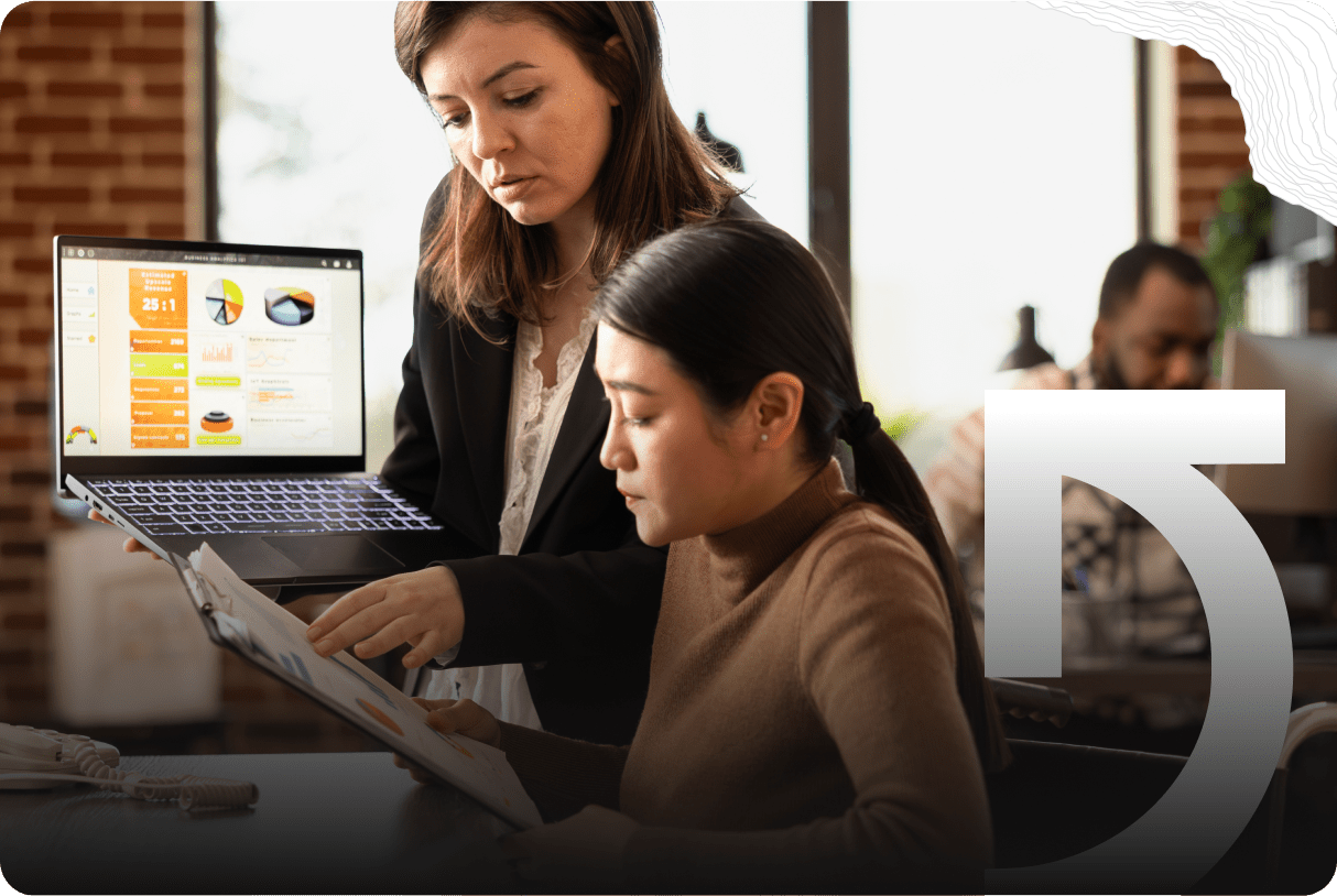 Two women collaborate at a desk, one holding a laptop displaying charts and graphs—visuals of their success stories—while the other reviews a document. Another person works in the background.